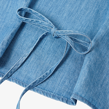 Macro product shot focusing on the fabric texture and construction of the tie-back ribbon detail on a denim blouse.