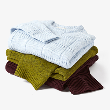 Creative fashion still life photography of a styled stack of folded knitwear sweaters in light blue, olive green, and deep burgundy on a crisp white background.